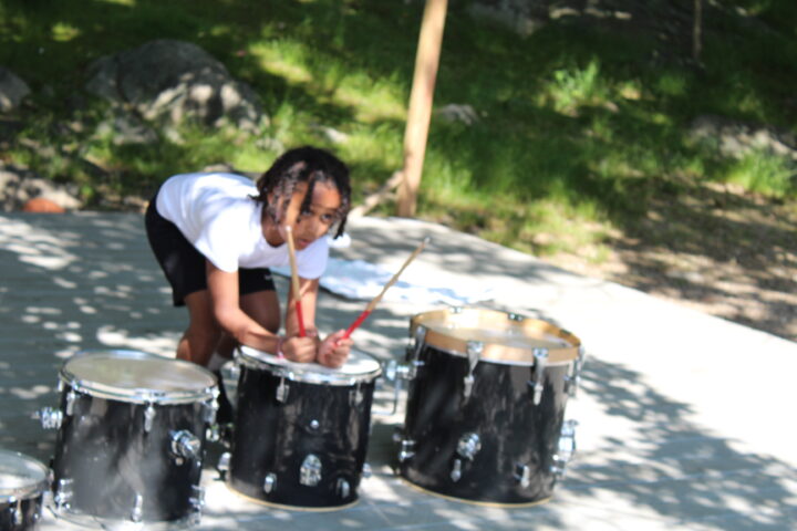 Smiling camper hitting 3 drums with drumsticks