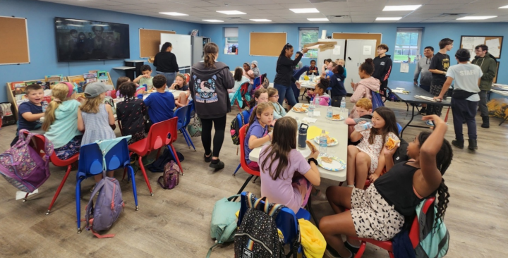 kids in a large conference room seated at tables in groups with several teachers standing and discussing